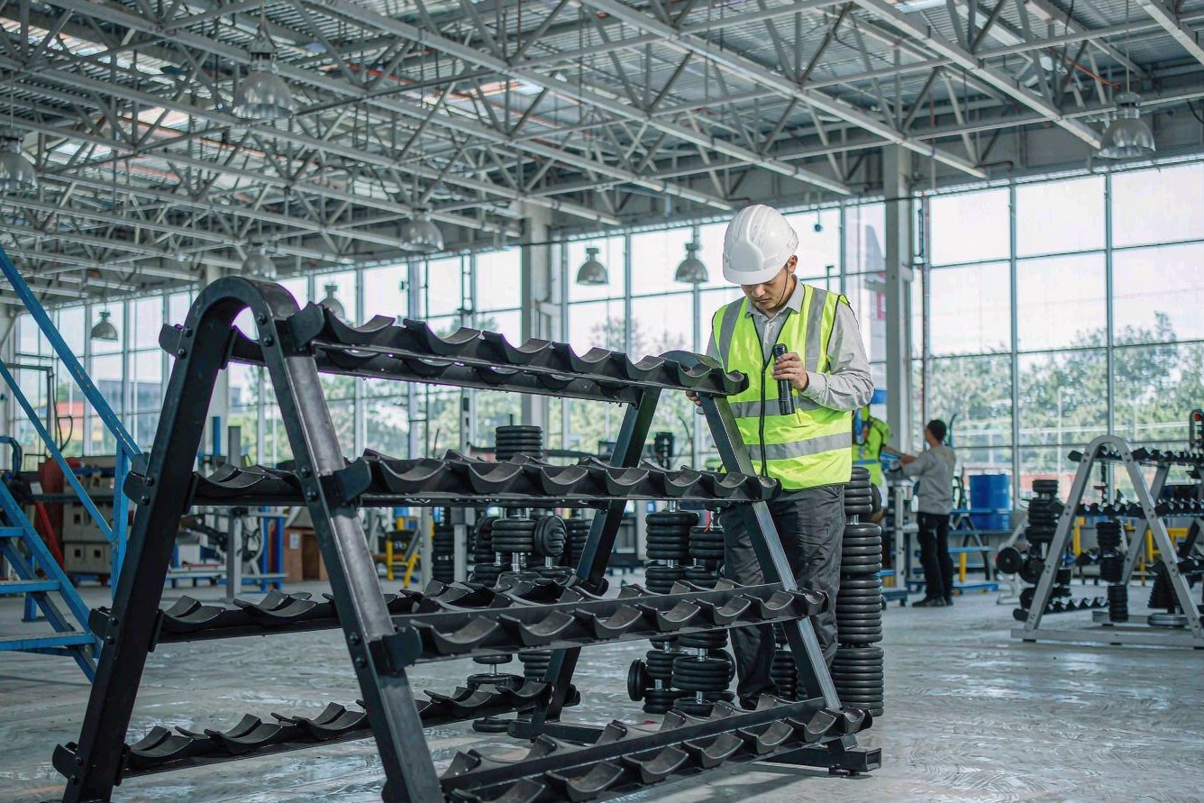 Factory auditor in a safety helmet inspecting a dumbbell rack production line with a flashlight